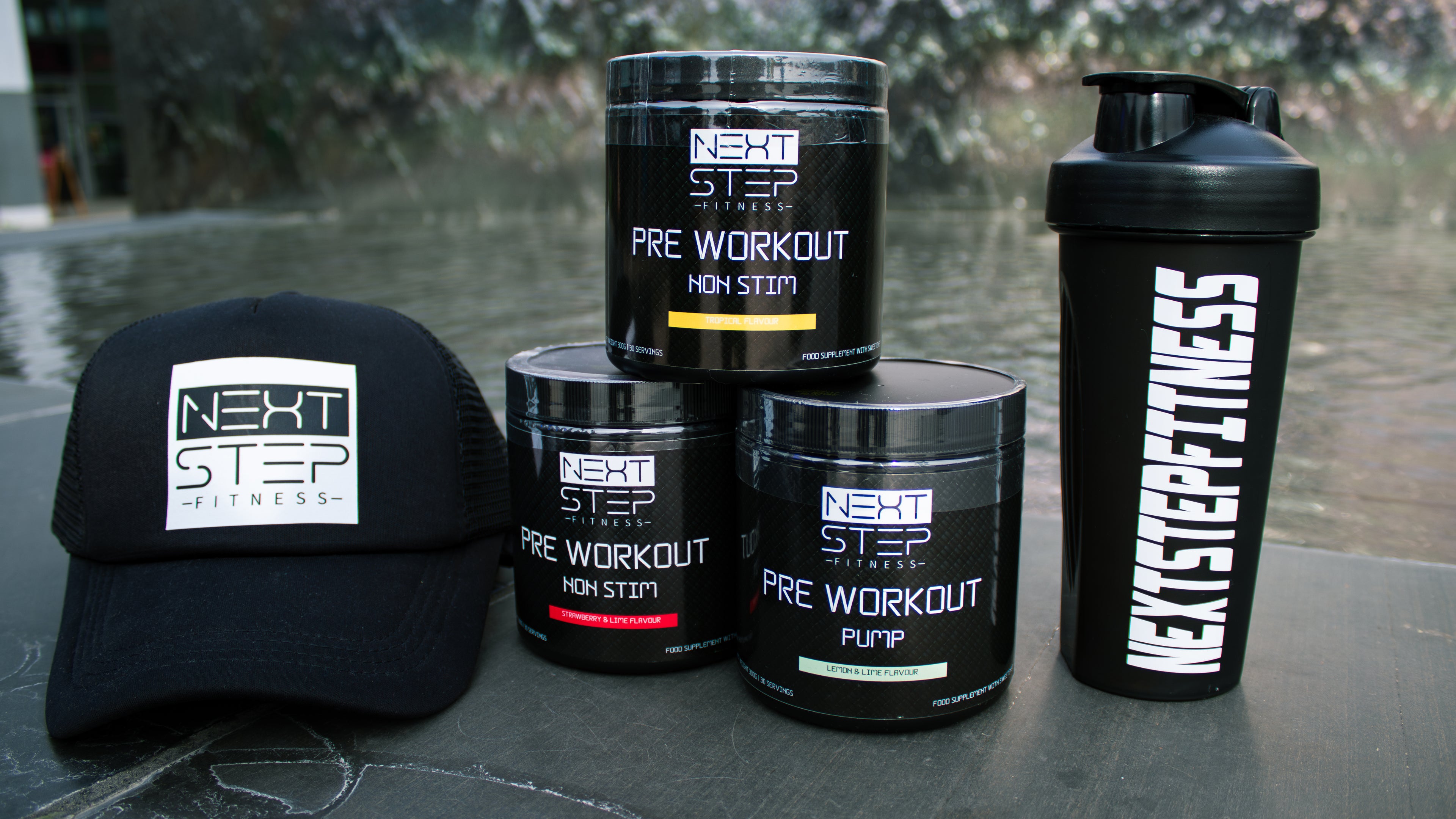 Collection of Next Step Fitness pre-workout products including a cap, shaker bottle, and containers on a textured surface.